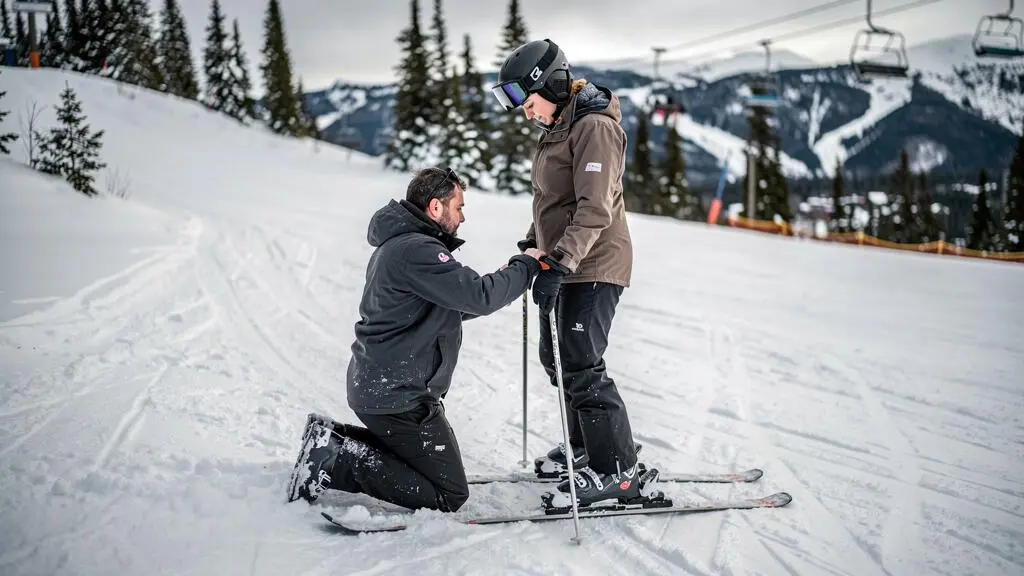 A ski instructor kneels beside an adult beginner on a gentle snowy slope at a Canadian resort, adjusting the beginner's skis and stance during a first lesson.