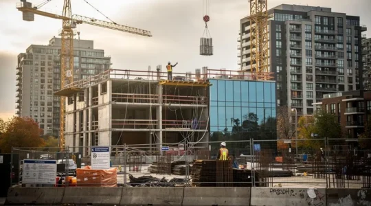 A construction worker on a high-rise residential building site in a Canadian city, with cranes and finished condo towers behind the site.