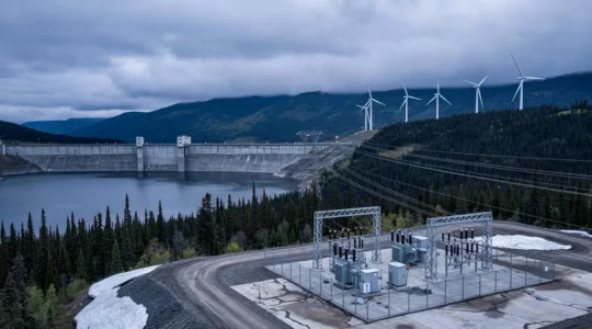 A hydroelectric dam with wind turbines and transmission lines visible across a forested Canadian landscape under an overcast sky.