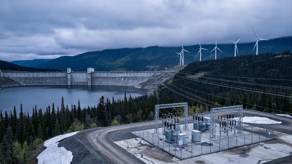 A hydroelectric dam with wind turbines and transmission lines visible across a forested Canadian landscape under an overcast sky.