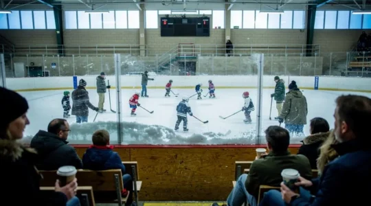Children skate during a community hockey practice at a small-town Canadian arena while families watch from the bleachers.
