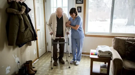 A nurse helping an elderly man walk with a walker in the hallway of his Canadian home.
