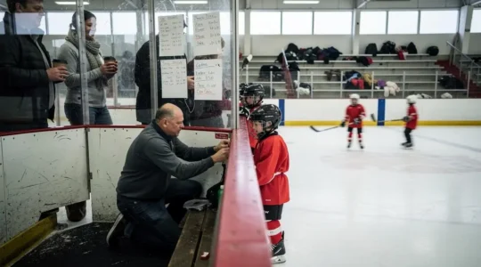 A volunteer coach kneels on a community arena bench adjusting a young hockey player's helmet while other children skate behind the boards.